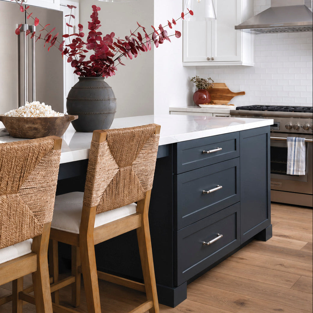 A modern kitchen island with dark navy blue cabinetry, a white countertop, and woven wood bar stools, set in a bright kitchen with wood floors and pendant lights.